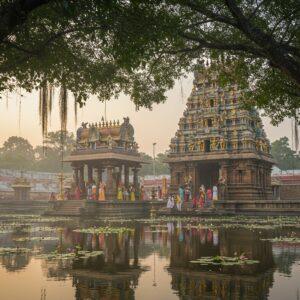 A majestic South Indian temple complex at dawn, its golden spire gleaming in the soft morning light. The ornate Gopuram towers above, adorned with intricate carvings of deities and mythological scenes in vibrant colors. In the foreground, a serene water tank reflects the temple's image, surrounded by blooming lotus flowers. Devotees in colorful traditional attire perform rituals near the entrance, offering flowers and lighting oil lamps. The scene is framed by ancient banyan trees with hanging roots, creating a mystical atmosphere. Soft mist lingers in the background, giving the scene an ethereal quality. The art style blends photorealistic details with subtle touches of digital enhancement, emphasizing the play of light and shadow on the temple's architecture. The color palette features rich golds, deep reds, and warm earth tones, balanced with cool blues in the sky and water.
