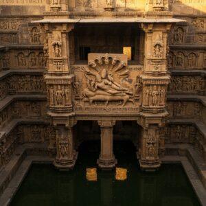 A majestic aerial view of Rani ki Vav stepwell in Gujarat, rendered in intricate detail with golden afternoon sunlight casting dramatic shadows across its multiple levels. The main focus is on a beautifully carved wall panel featuring a detailed sculpture of Lord Vishnu reclining on Shesha Naga, carved in warm sandstone. Surrounding this central piece are intricate geometric patterns, smaller sculptures of apsaras in graceful dance poses, and mythological scenes carved with incredible detail. The stepwell's architecture follows perfect geometric symmetry with seven levels visible, each adorned with over 500 principal sculptures and over 1000 minor ones. Water at the bottom reflects the intricate architecture, creating a mirror effect. The scene is enhanced with subtle mystical elements like soft golden halos around the sculptures and ethereal mist rising from the water. The art style combines hyperrealistic architectural detail with traditional Indian sculptural aesthetics, featuring a color palette of warm sandstone, deep gold, and cool water tones. The lighting is dramatic, highlighting the three-dimensional quality of the sculptures while maintaining the spiritual atmosphere of this ancient architectural marvel.