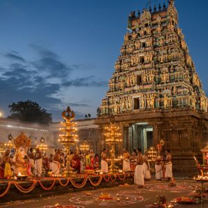 A majestic night scene of a grand South Indian Shiva temple with intricate Dravidian architecture, illuminated by hundreds of traditional brass oil lamps and modern spotlights. The main gopuram towers 7 tiers high, adorned with detailed sculptures of Hindu deities in gold and vibrant colors. In the foreground, devotees in traditional attire perform aarti with gleaming brass lamps, creating circles of light. Lord Shiva's sacred bull Nandi, detailed in polished black stone, faces the sanctum. Multiple festival elements are artistically arranged: colorful kolam patterns, strings of marigold and jasmine flowers, and floating lotus flowers in the temple tank. The scene includes both traditional elements (priests in white dhotis performing rituals) and modern touches (subtle LED lighting). The atmosphere is enhanced by swirling incense smoke, creating a mystical ambiance. The color palette combines deep blues of twilight sky, warm golden temple lights, saffron and red ritual decorations, with ethereal silver moonlight casting shadows. The style merges photorealistic temple architecture with ethereal Madhubani-inspired patterns in the sky and surrounding elements.