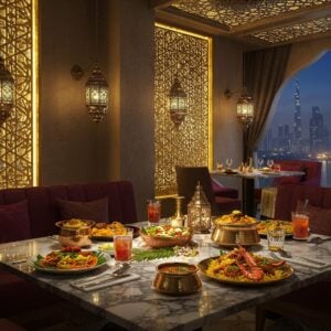 A luxurious modern restaurant interior with geometric Islamic patterns on golden-lit walls, merging with traditional Kerala architectural elements. In the foreground, a stunning marble dining table adorned with brass and copper traditional Kerala serving vessels. The table features an elegant spread of Kerala-Arabic fusion dishes - including mandi rice garnished with Kerala spices, grilled prawns with Arabian herbs, and saffron-infused biriyani. Soft ambient lighting from ornate Arabian lanterns creates a warm golden glow. The scene includes intricate mandala patterns in peacock blue and gold on the ceiling. Contemporary minimalist seating with plush velvet cushions in deep maroon. Crystal glasses filled with traditional drinks add sparkle. In the background, a large window reveals a dramatic Dubai skyline at dusk, with Burj Khalifa visible. The overall composition combines luxury fine dining aesthetics with cultural elements from both Kerala and Arab traditions. The scene is rendered in a photorealistic style with cinematic lighting, emphasizing the fusion of cultures through food and architecture.
