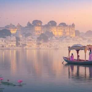 An ethereal scene of Udaipur at sunrise, where the tranquil waters of Lake Pichola reflect the vibrant hues of the sky. In the foreground, an intricately detailed boat adorned with traditional Indian motifs floats gracefully, guided by a serene Indian woman in a modern, flowing pastel saree. The majestic City Palace rises in the background, its architecture a blend of ancient grandeur and modern elegance. Soft, glowing lights illuminate the palace and surrounding temples, highlighting their spiritual essence. Lotus flowers float on the lake, symbolizing purity and enlightenment. The overall ambiance is calm and inviting, with a harmonious balance of tradition and contemporary aesthetics. The art style is serene and spiritual, with pastel colors and gentle lighting that evoke a sense of mindfulness and peace.