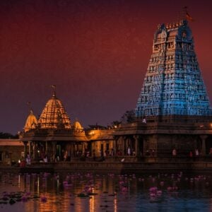 A serene depiction of the Chengannur Mahadeva Temple, set against a twilight sky that blends hues of deep maroon and peacock blue, with soft glowing lights illuminating the temple's intricate carvings and ornate architecture. The temple is nestled amidst lush greenery, with lotus flowers gently floating on a tranquil water surface in the foreground. Silhouettes of devotees are seen performing traditional rituals, their figures softly highlighted by the warm glow of temple lamps. A large, ethereal mandala subtly overlays the scene, symbolizing spiritual harmony and mindfulness. The image blends traditional Indian art aesthetics with modern digital techniques, creating a visually captivating and culturally respectful representation of this divine destination.