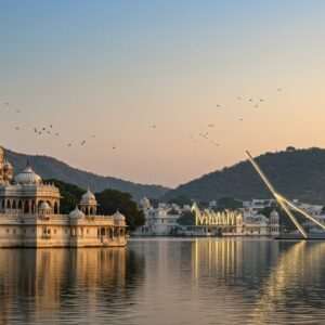 A mesmerizing scene of Udaipur's cityscape blending tradition and modernity, with the iconic Lake Pichola in the foreground. On one side, the majestic Jagdish Temple stands proudly, its intricate carvings and towering spires highlighted by the warm glow of sunset. On the other side, a modern art installation with sleek lines and glowing elements adds a contemporary touch. The serene waters of the lake reflect both the ancient and the new, creating a harmonious balance. In the background, the Aravalli hills provide a tranquil backdrop. The sky is painted with hues of soft blue, pale pink, and warm gold, casting a peaceful ambiance over the scene. This ethereal image captures the essence of a spiritual journey in Udaipur, inviting exploration and discovery. The art style combines traditional Indian motifs with modern digital aesthetics for a captivating visual experience.