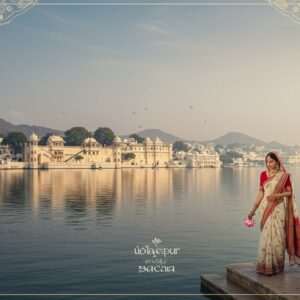A breathtaking panoramic view of Udaipur, showcasing the serene waters of Lake Pichola reflecting the majestic City Palace. In the foreground, an elegantly dressed modern Indian woman in a contemporary saree walks gracefully along the lakeside, embodying the essence of a modern pilgrim. She carries a delicate lotus flower, symbolizing purity and spirituality. The scene is bathed in soft, ethereal light, with hues of pastel blues and warm golds enveloping the horizon. Traditional elements like intricate patterns of peacock motifs and mandalas subtly blend into the modern digital aesthetics, creating a harmonious fusion of tradition and contemporary elegance. The overall ambiance is tranquil and inviting, drawing the viewer into Udaipur's timeless beauty.