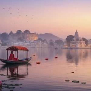 A breathtaking scene of Udaipur's serene Lake Pichola at dawn, with the majestic City Palace in the background. The sky is painted with soft pastel hues of pink, lavender, and gold, reflecting on the calm waters. A traditional Rajasthani boat glides gently across the lake, carrying a serene Indian woman dressed in an elegant saree, her hands folded in prayer. In the distance, the silhouette of the Jag Mandir temple is visible, adorned with glowing diyas (oil lamps) that cast a warm, spiritual glow. Large lotus flowers float on the water's surface, adding to the ethereal ambiance. The scene captures a perfect blend of spirituality and the cultural richness of Udaipur, with a modern digital aesthetic emphasizing tranquility and devotion.