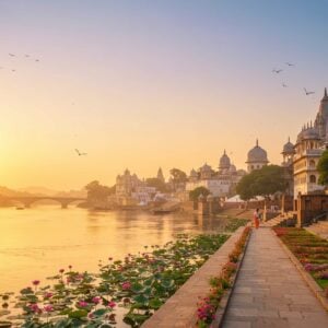 A breathtaking scene of the ancient city of Hastinapura, blending historical grandeur with modern aesthetics. The foreground features a serene landscape with the Ganges River flowing gently, reflecting the soft glow of a warm golden sunrise. In the background, majestic palaces and temples of Hastinapura rise against a sky painted in pastel hues of pink and blue. Intricate architectural details are adorned with motifs inspired by traditional Indian art forms like Madhubani and Pattachitra. Elegant pathways, lined with blooming lotus flowers and lush greenery, lead towards the city, inviting exploration. The scene is tranquil yet vibrant, capturing the essence of both heritage and contemporary allure.