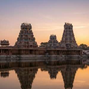 A breathtaking panorama showcasing an ancient Indian temple complex under a serene, pastel-colored sky with soft hues of dawn. The temple structures are intricately carved with traditional motifs like lotus flowers, mandalas, and celestial beings, echoing the grandeur of Chola or Dravidian architecture. The temple walls are adorned with rich colors of maroon, saffron, and gold, highlighted by delicate digital enhancements that give a modern touch. In the foreground, a peaceful reflecting pond mirrors the temple, creating a tranquil and harmonious atmosphere. The scene is imbued with a soft, ethereal glow that accentuates the spiritual essence and divine symbolism inherent in the architecture. The overall composition feels sacred and inviting, drawing the viewer into a journey of cultural and architectural discovery.
