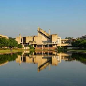 A sprawling view of Chandigarh's Capitol Complex, showcasing the blend of modernist architecture and lush greenery. The image focuses on Le Corbusier's iconic structures like the Secretariat, High Court, and Assembly, with clean lines and geometric shapes. The buildings are depicted in their original, earthy tones contrasted against a clear blue sky. In the foreground, a reflection of these structures on a tranquil water body adds depth and harmony to the scene. The art style should be hyperrealistic, capturing the essence of mid-20th-century modernism with a touch of digital finesse, highlighting the balance between urban planning and nature. The lighting is soft and natural, emphasizing the serene yet innovative design of Chandigarh's architecture.