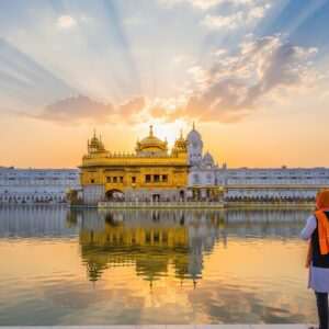 A serene and ethereal depiction of the Golden Temple in Amritsar, bathed in the warm glow of a setting sun. The temple's golden facade reflects brilliantly in the calm, sacred waters surrounding it. In the foreground, an Indian pilgrim dressed in a traditional yet modern fusion attire stands in awe, hands folded in reverence. Soft, pastel-colored clouds drift in the sky above, with gentle rays of light highlighting the temple's intricate details. The scene captures the tranquility and spiritual significance of this sacred site, blending modern digital aesthetics with rich cultural heritage. The atmosphere exudes peace, devotion, and a sense of timeless beauty.