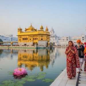 An enchanting depiction of the Golden Temple in Amritsar, its golden dome reflecting in the calm, serene waters surrounding it. The scene captures the early morning light, casting a soft, ethereal glow over the temple and its surroundings. In the foreground, a group of Indian pilgrims, dressed in traditional attire with intricate patterns reminiscent of Madhubani art, walk along Heritage Street with expressions of awe and devotion. The street is lined with ornate architecture, blending historical elements with modern aesthetics. The atmosphere is tranquil and spiritual, with pastel hues of soft blue and warm gold highlighting the scene's sacredness. Large lotus flowers float gently on the water surface, enhancing the surreal and spiritual tone of the image.