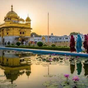 A majestic depiction of the State Gurudwara in Kapurthala, bathed in the warm glow of a setting sun. The architecture is intricately detailed with elements of traditional Sikh design, highlighted by golden domes and ornate carvings. In the foreground, a serene pond reflects the structure, surrounded by lush greenery and blooming lotus flowers. A group of Indian pilgrims, dressed in traditional attire with vibrant turbans and dupattas, walk towards the Gurudwara with a sense of devotion and peace. The scene is ethereal and tranquil, capturing the spiritual essence and historical significance of this iconic landmark. The use of rich colors like gold, maroon, and peacock blue adds depth and vibrancy to the image, making it both modern and timeless.