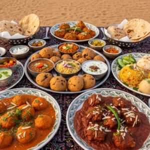 A vibrant scene depicting a traditional Rajasthani feast laid out on an intricately patterned Phad-style tablecloth. The table is adorned with a variety of colorful dishes such as dal baati churma, gatte ki sabzi, and laal maas, each exquisitely garnished with fresh herbs and spices. In the background, a serene desert landscape with soft pastel hues of sunset casts a warm glow over the scene. Traditional Indian crockery with detailed Pattachitra designs holds the food. The ambiance is enhanced by ethereal golden lights, highlighting the rich cultural motifs like mandalas and peacock feathers subtly integrated into the setting. The overall style is hyperrealistic with a modern twist, emphasizing both the heritage and innovation of Rajasthan's culinary art.