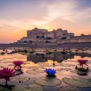 An awe-inspiring view of the majestic Chittorgarh Fort bathed in the golden glow of the setting sun, casting long shadows on its ancient walls. The scene captures the grandeur of the fort's architecture with its towering battlements and intricate stone carvings. In the foreground, vibrant maroon and peacock blue lotus flowers float gently on a serene water surface, reflecting the fort's image. A soft, ethereal light envelops the scene, adding a mystical aura. The sky is painted in pastel hues of pink and warm gold, dotted with wispy clouds. The overall atmosphere is one of tranquility and timeless beauty, inviting viewers to explore the fort's rich history and mysteries.