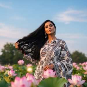A serene and ethereal scene depicting a beautiful Indian woman with glossy, flowing black hair, free of flakes, standing in a tranquil garden surrounded by softly glowing lotus flowers. The background features a pastel sky with gentle hues of soft blue and pale pink, illuminated by warm golden light. She is dressed in a modern fusion of traditional and contemporary attire, with subtle intricate patterns inspired by Madhubani art on her outfit. Her expression exudes confidence and serenity, symbolizing freedom from dandruff. The overall atmosphere is calm and inviting, with an emphasis on purity and rejuvenation, capturing the essence of Pilgrim's anti-dandruff range.