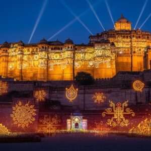 A majestic depiction of Chittorgarh Fort at twilight, illuminated by a captivating sound and light show. The fort's towering stone structures are bathed in a warm golden glow, with intricate architectural details highlighted against the deepening indigo sky. Ethereal, soft glowing lights dance across the ancient walls, creating a surreal atmosphere. In the foreground, traditional Indian motifs and cultural symbols subtly blend with the scene, enhancing its historical essence. The lighting is dynamic and dramatic, with vibrant hues of saffron, maroon, and peacock blue adding depth and allure. Art style is hyperrealistic with a touch of digital fantasy, capturing the grandeur and mystique of this iconic heritage site.