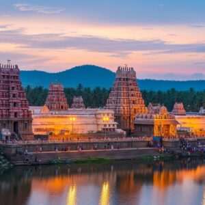 A serene, ethereal depiction of the ancient Tulu Nadu temples, nestled amidst lush green landscapes and rolling hills under a soft pastel sky. The temples are intricately designed with traditional architectural elements, featuring ornate carvings and sculptures, adorned with vibrant maroon and gold accents. A gentle, warm glow illuminates the temple surroundings, highlighting the serene ambiance. In the foreground, a peaceful river reflects the temples and the soft glow of the setting sun. Pilgrims in traditional Indian attire are seen respectfully approaching the temples, creating a scene of devotion and tranquility. The atmosphere is calm and spiritual, capturing the essence of a sacred journey.