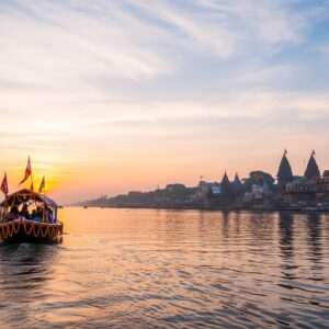 A serene and ethereal scene of the Hooghly River merging with the Ganga, depicted at sunrise. The river shimmers with a soft golden hue as the first light of dawn kisses its surface. In the foreground, a traditional Indian boat drifts gently on the calm waters, adorned with vibrant marigold garlands and fluttering flags. Along the riverbanks, lush greenery and ancient temple silhouettes evoke a sense of timeless spirituality. In the sky, soft pastel shades of pink and blue blend seamlessly, creating an atmosphere of tranquility and reverence. The overall composition exudes a hyperrealistic and surreal quality, inviting viewers into the sacred connection between the Hooghly and the Ganga.