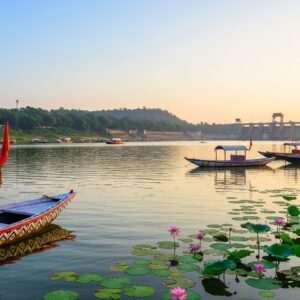 A serene and ethereal landscape of the Kanhar River at sunrise, with soft glowing lights reflecting off the calm water surface. In the foreground, traditional Indian boats with intricate patterns float gently, while lush greenery and blooming lotus flowers line the riverbanks. In the distance, a modern dam stands majestically, blending seamlessly with the natural surroundings. The sky is painted in pastel hues of soft blue and pale pink, casting a warm golden glow over the scene. The art style is hyperrealistic, capturing the tranquil beauty and significance of the river in a harmonious blend of tradition and modernity.