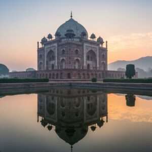 A serene and ethereal image of the ancient town of Sasaram at dawn, featuring the majestic tomb of Sher Shah Suri, bathed in a soft, golden light. The tomb, an epitome of Indo-Islamic architecture, stands tall with its intricate stone carvings and domed structure. In the foreground, a calm water body reflects the monument, surrounded by lush greenery. The sky is painted in pastel hues of soft blue and pale pink, with a gentle mist adding a surreal quality. In the background, the silhouette of distant hills creates a sense of depth and tranquility. The overall tone is one of spiritual serenity and historical grandeur, inviting viewers into a journey of discovery and reflection.